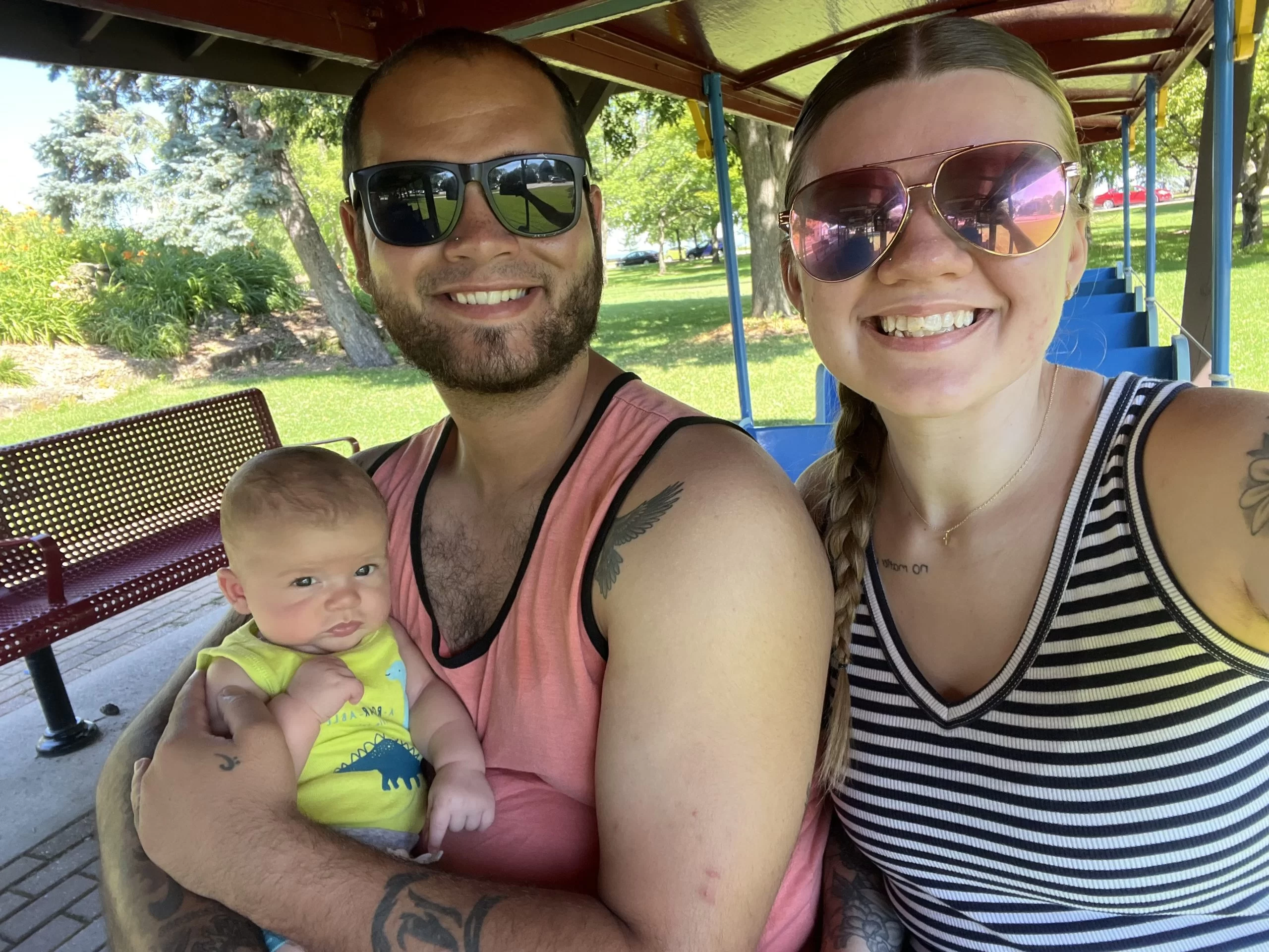 Woman and Man outside under a picnic shelter, both wearning sunglasses holding an adorable small baby