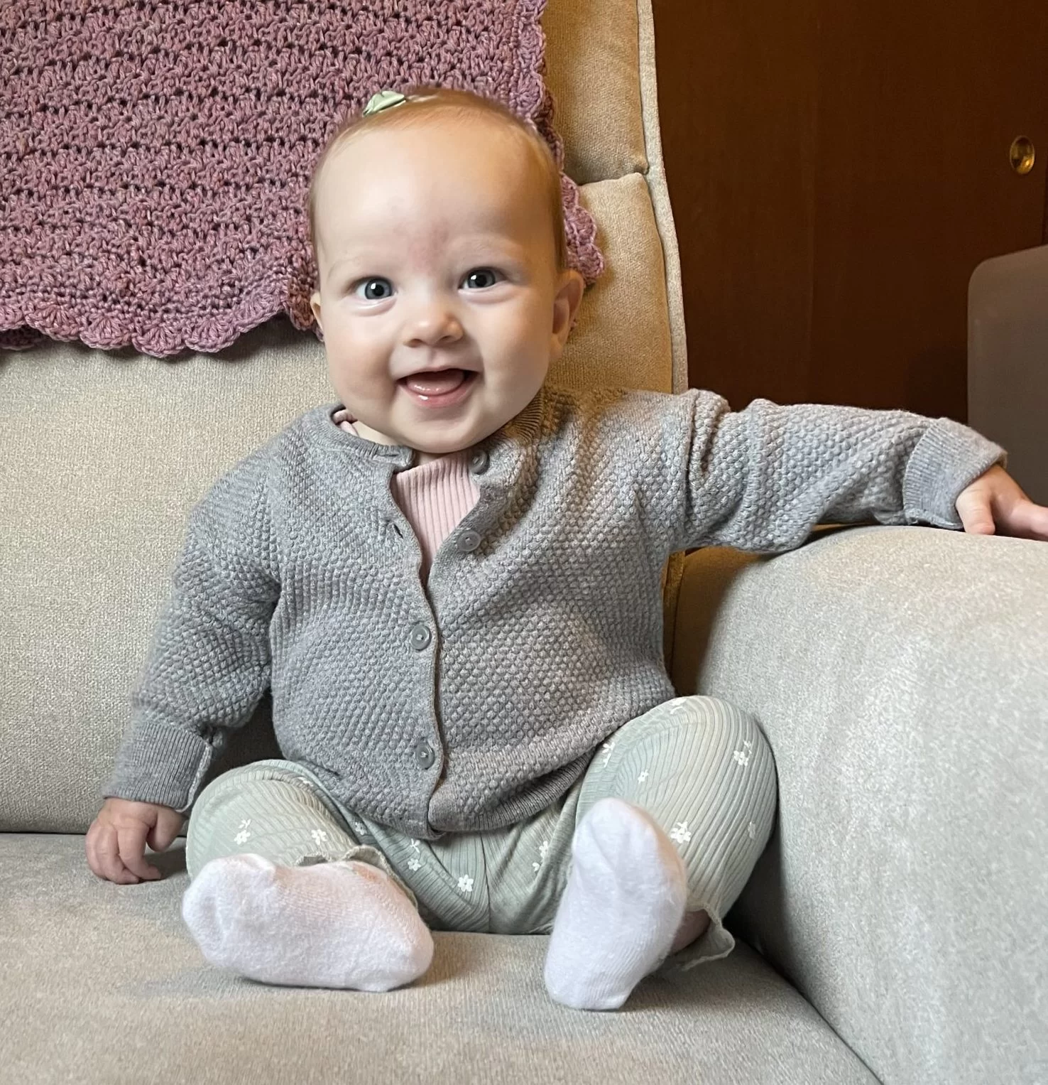 a smiling baby in a gray sweater happy after donating breast milk in a tan upholstered recliner with a mauve blanket behind her.