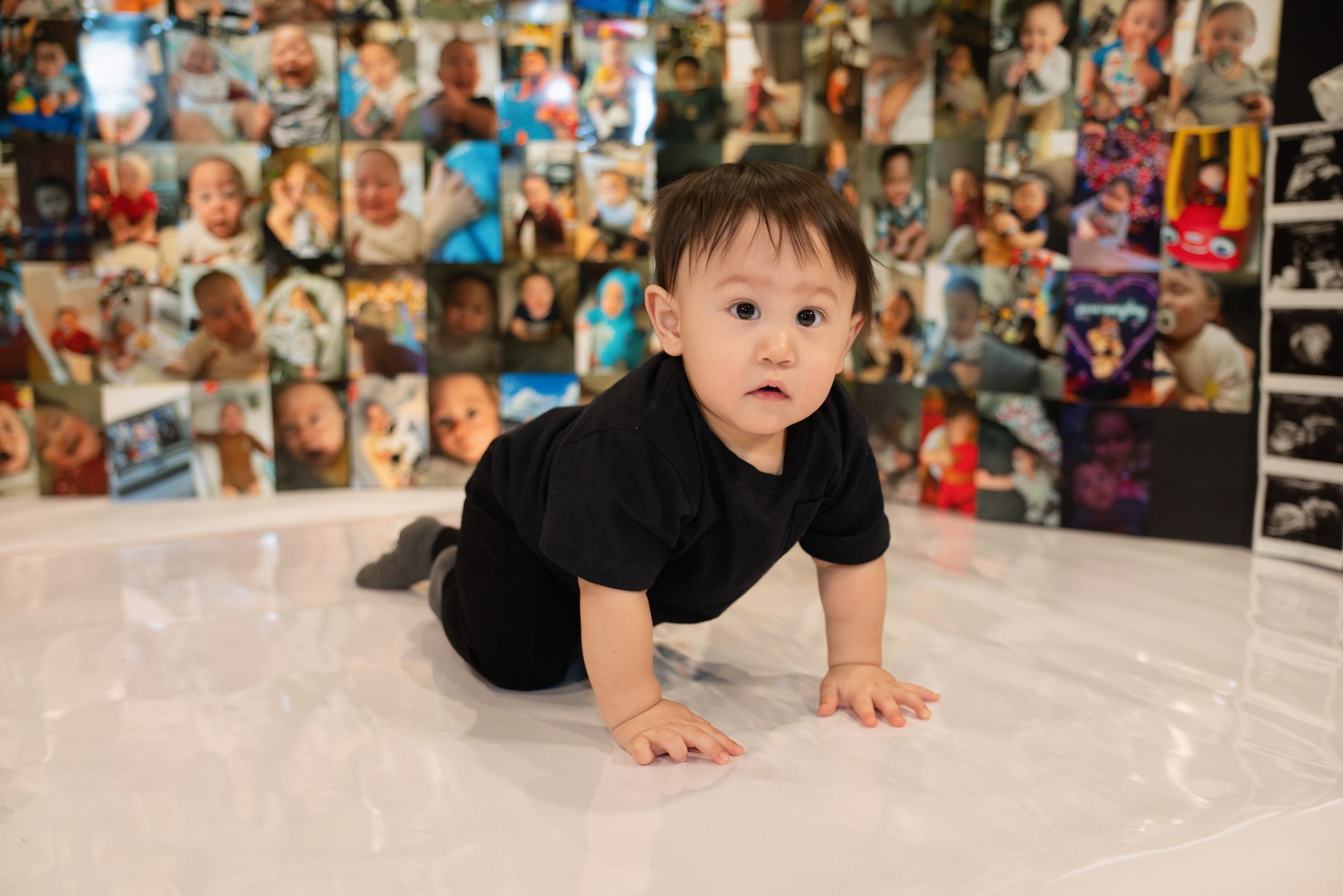 A year of growth for healthy baby jet in front of all his baby pictures in a black shirt