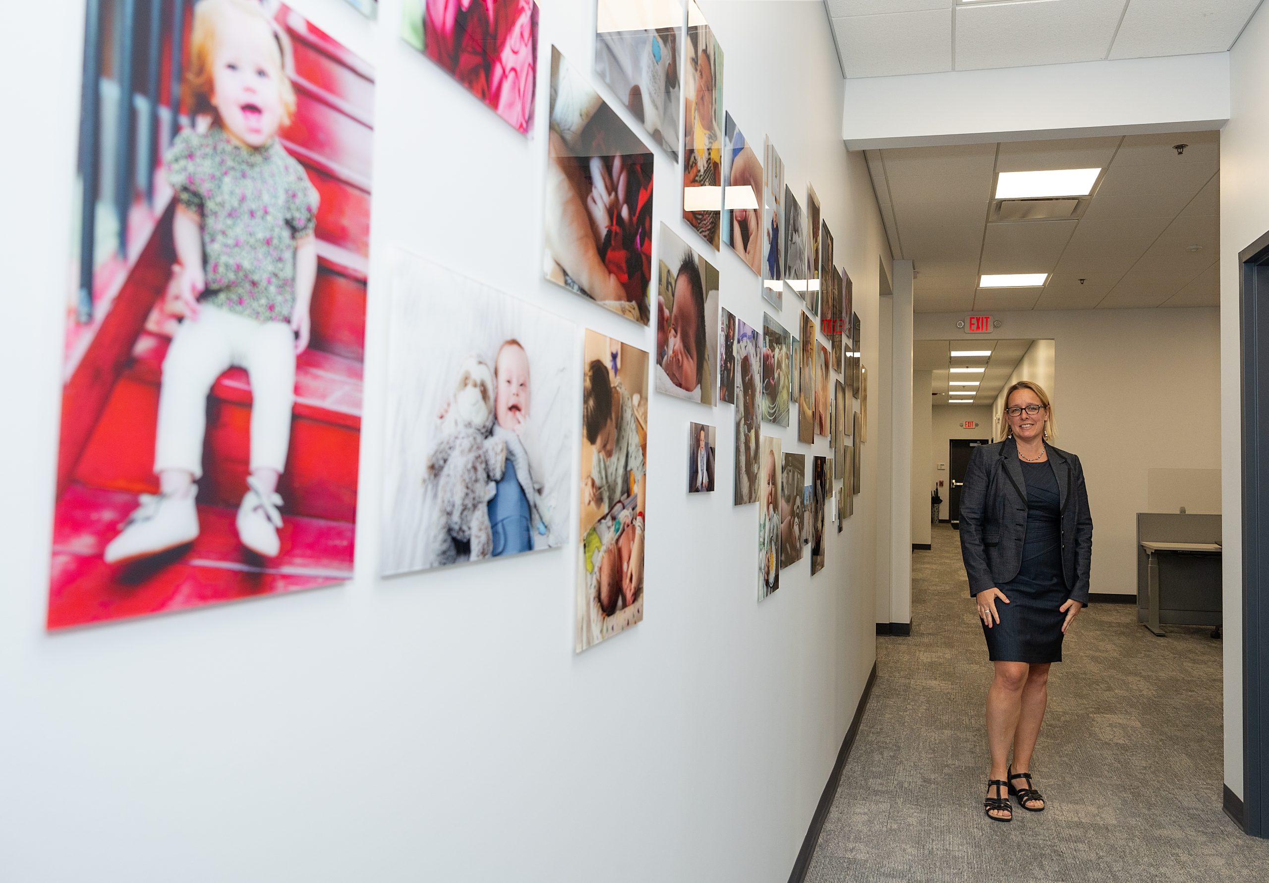 Blonde woman in dark pant suit stands in the back of a row of baby photos down an office hallway